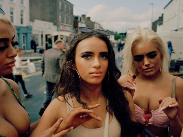 Three young women pose in an urban setting. The woman in the center wears sunglasses on her head and a light-colored tank top.