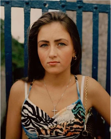 Portrait of a young woman wearing a patterned top and a cross necklace in front of a blue metal fence.