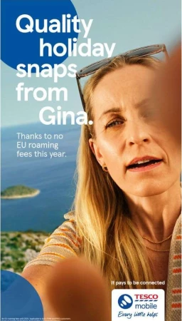 An adult woman with long blonde hair is taking a selfie on vacation against a coastal backdrop for a Tesco Mobile advertisement with clear text.