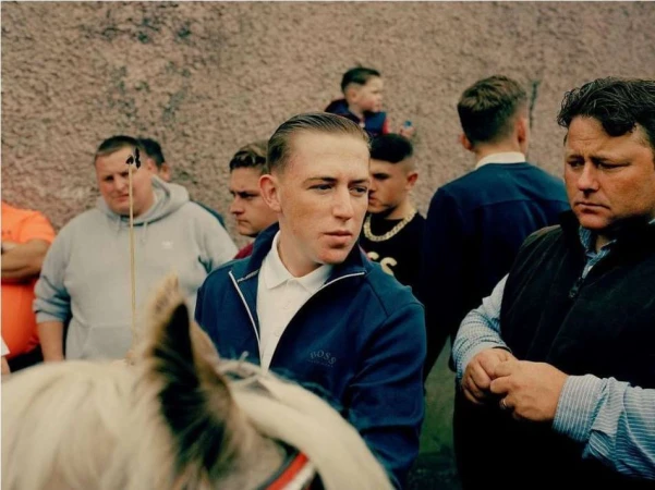 Crowd of people gathered outdoors near a horse; men are wearing casual clothing, in a medium shot with a textured background.