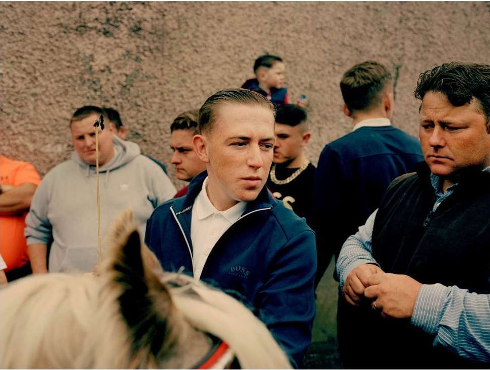 A crowd of people, including an adult male in a blue jacket, stand together in an outdoor setting on what appears to be a cloudy day.