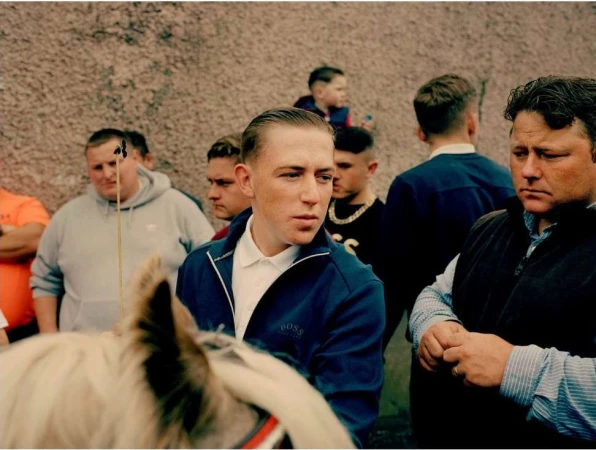 A crowd of people, including an adult male in a blue jacket, stand together in an outdoor setting on what appears to be a cloudy day.