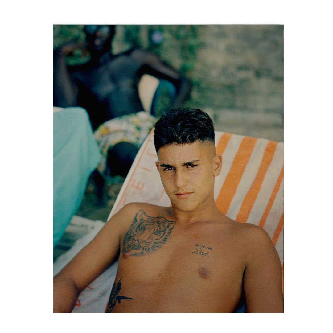 Young man with chest tattoos lounges on a striped beach chair outdoors, with a blurred person sitting in the background.