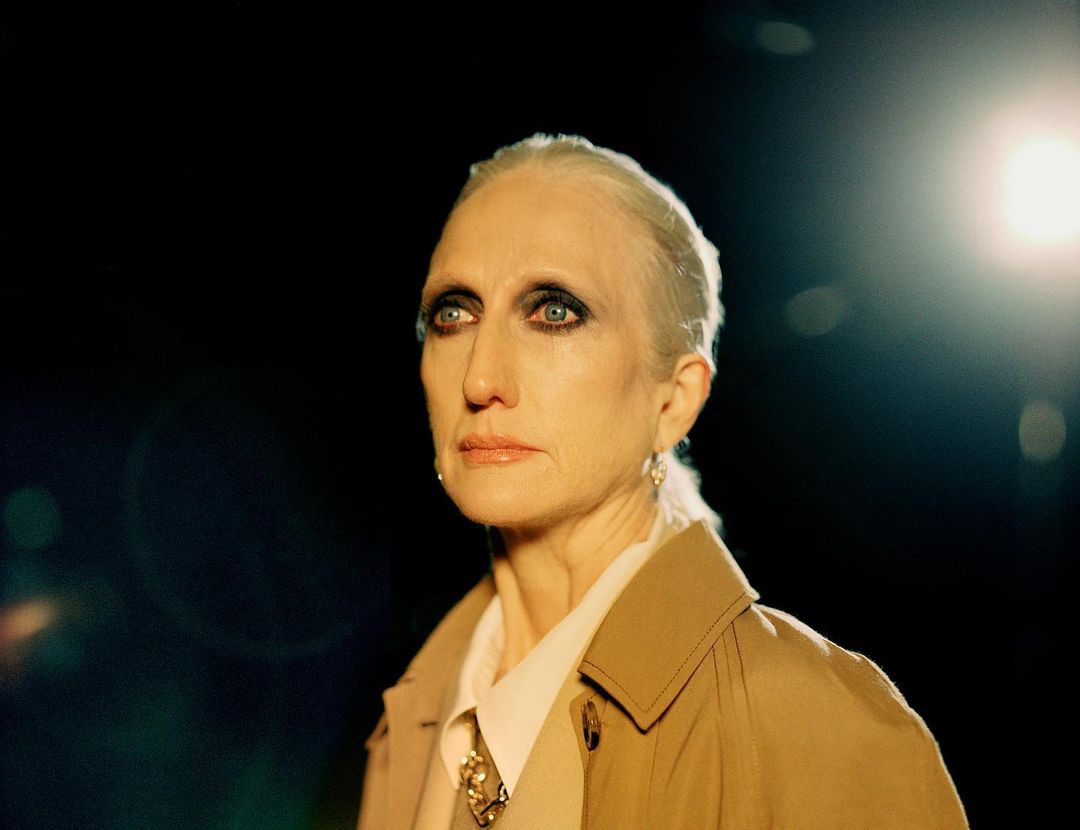 Close-up studio shot of a mature adult woman with white hair wearing a trench coat, looking off-camera against a dark background.