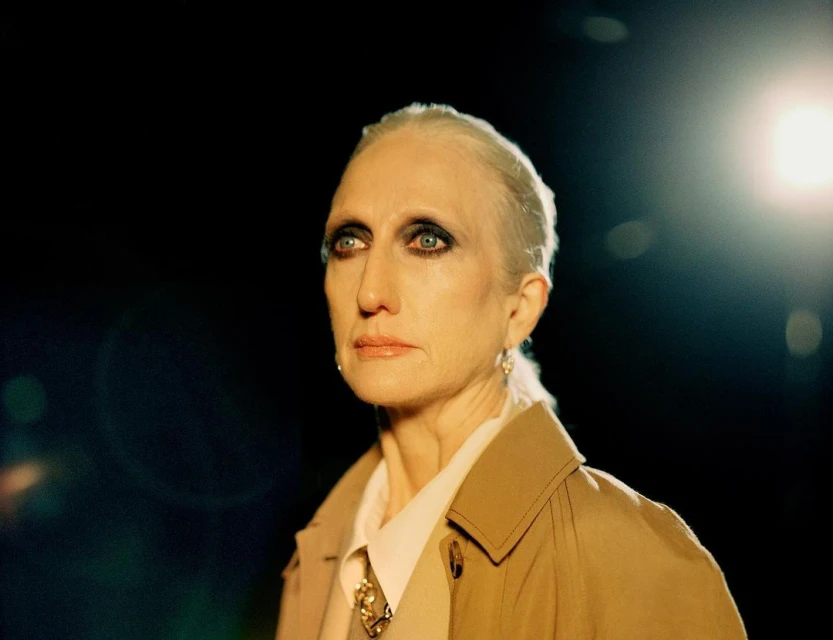 Close-up studio shot of a mature adult woman with white hair wearing a trench coat, looking off-camera against a dark background.