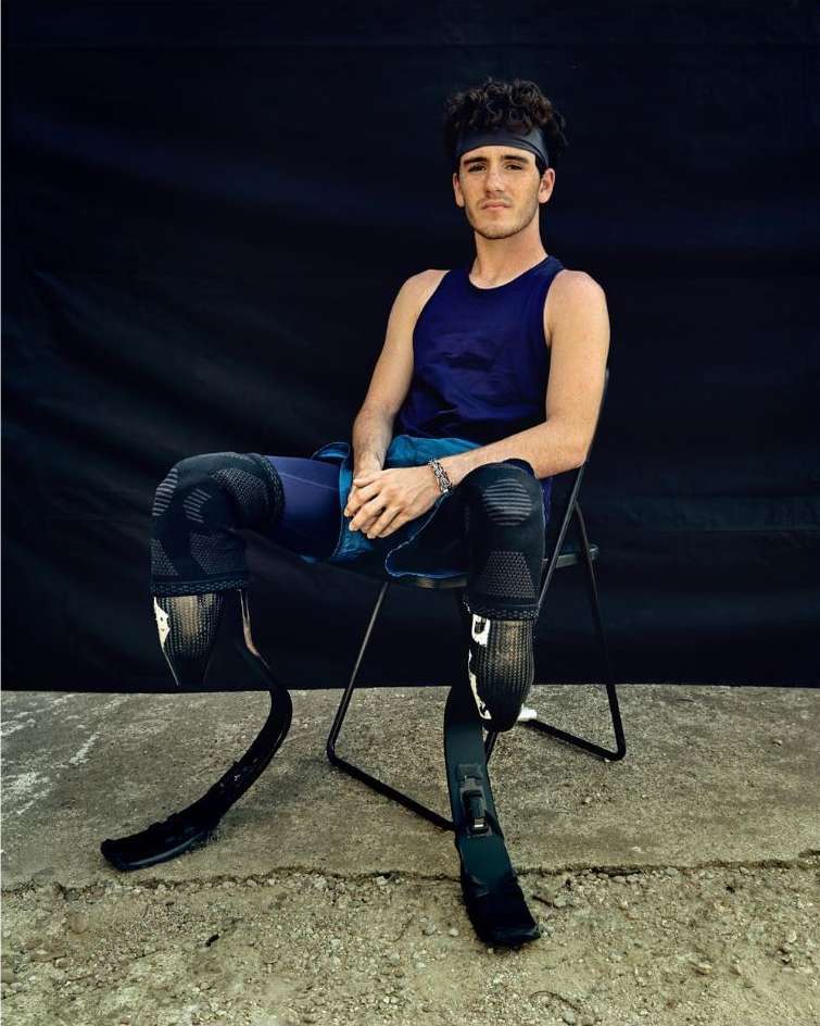 Athletic man with prosthetic legs poses seated against a dark backdrop; he wears sportswear, a headband and a bracelet in a natural light shot.