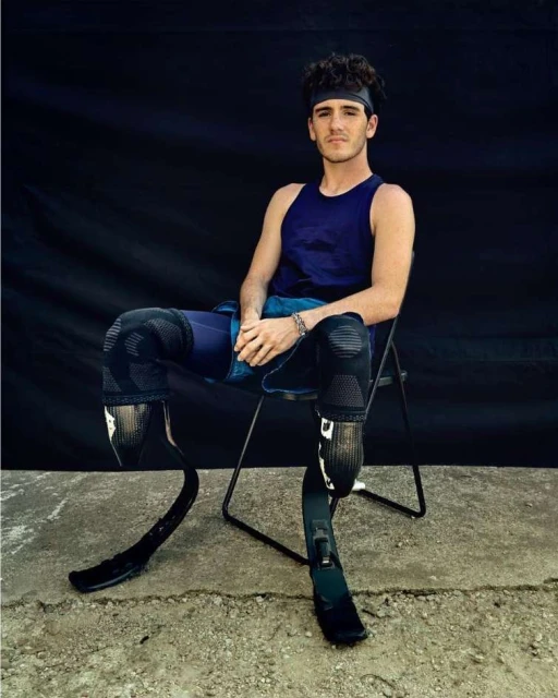 Athletic man with prosthetic legs poses seated against a dark backdrop; he wears sportswear, a headband and a bracelet in a natural light shot.