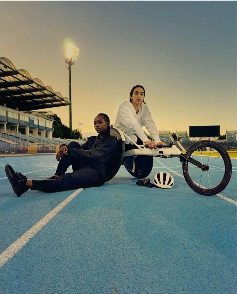 Two female athletes with a handcycle on a blue track field in stadium seating during daytime, under an overcast sky.