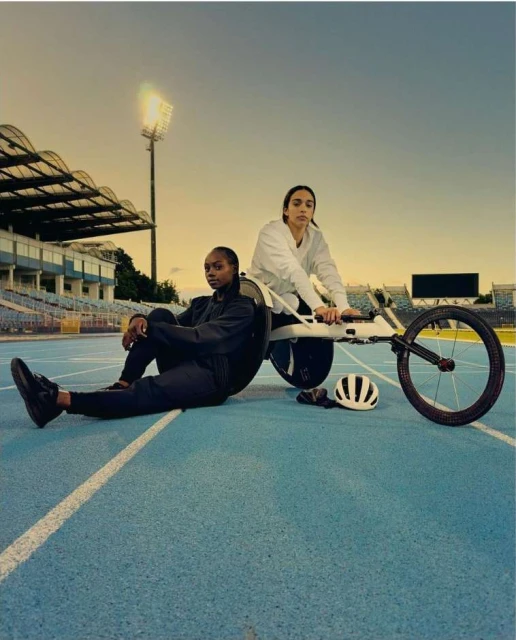 Two female athletes with a handcycle on a blue track field in stadium seating during daytime, under an overcast sky.