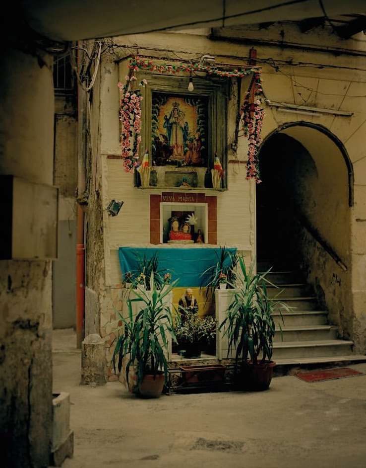 Exterior view of a shrine in a weathered alleyway featuring religious icons, potted plants, a blue cloth, framed artwork, and stairs.