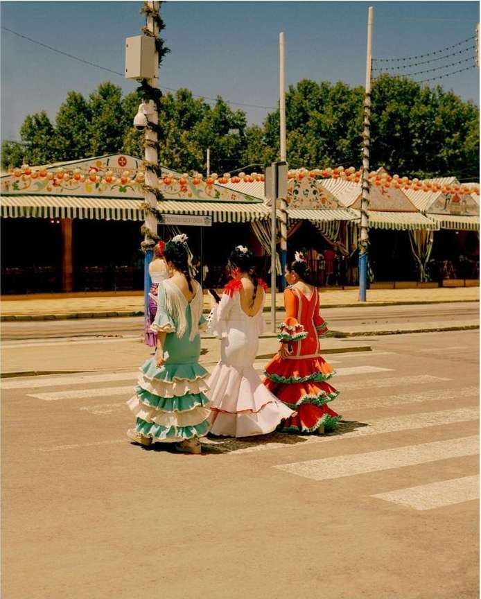 Three women in flamenco dresses crossing a street with white crosswalk lines, buildings and trees in the background on a sunny day.