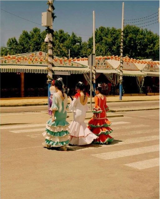 Three women in flamenco dresses crossing a street with white crosswalk lines, buildings and trees in the background on a sunny day.