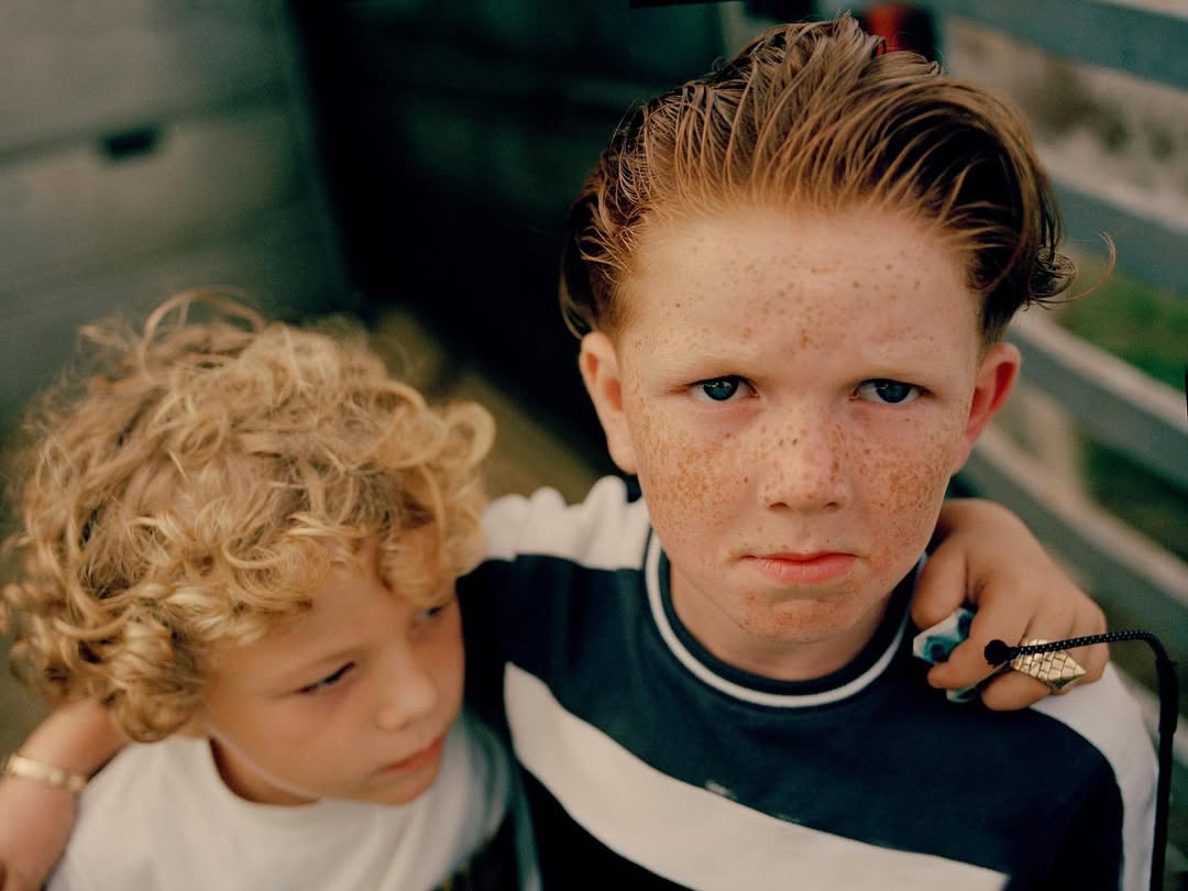 Close up of two young boys, one with red hair and freckles, the other with blond curly hair, standing close together in an indoor setting.