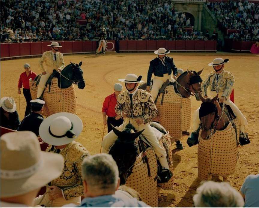 Bullfighters on horses in full regalia are positioned in a sandy arena, with a densely packed crowd visible in the background.