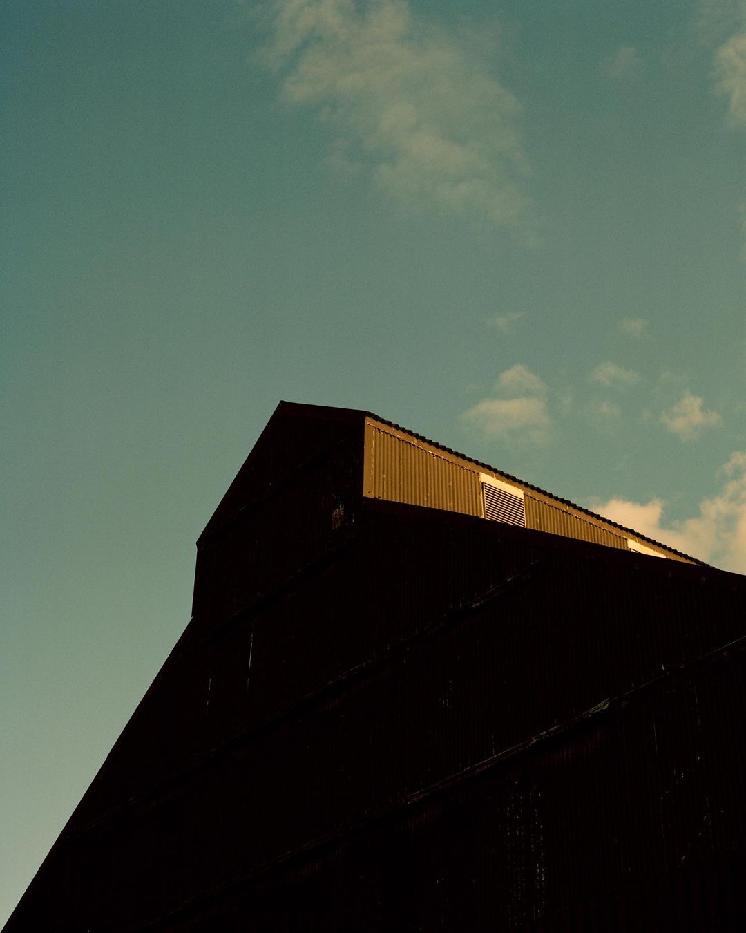 Low angle of a weathered, gable-roofed building against a pale blue sky with scattered clouds, capturing architectural texture.