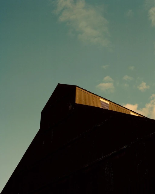 Low angle of a weathered, gable-roofed building against a pale blue sky with scattered clouds, capturing architectural texture.