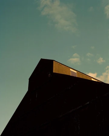 Low angle of a weathered, gable-roofed building against a pale blue sky with scattered clouds, capturing architectural texture.
