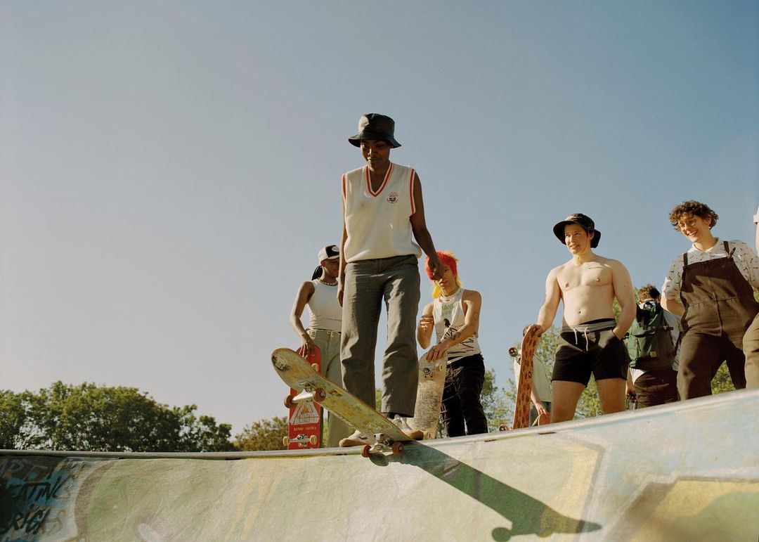 Teenager in bucket hat skateboarding in an outdoor skate park with a group of people watching on a sunny day.