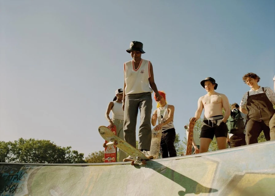 Teenager in bucket hat skateboarding in an outdoor skate park with a group of people watching on a sunny day.