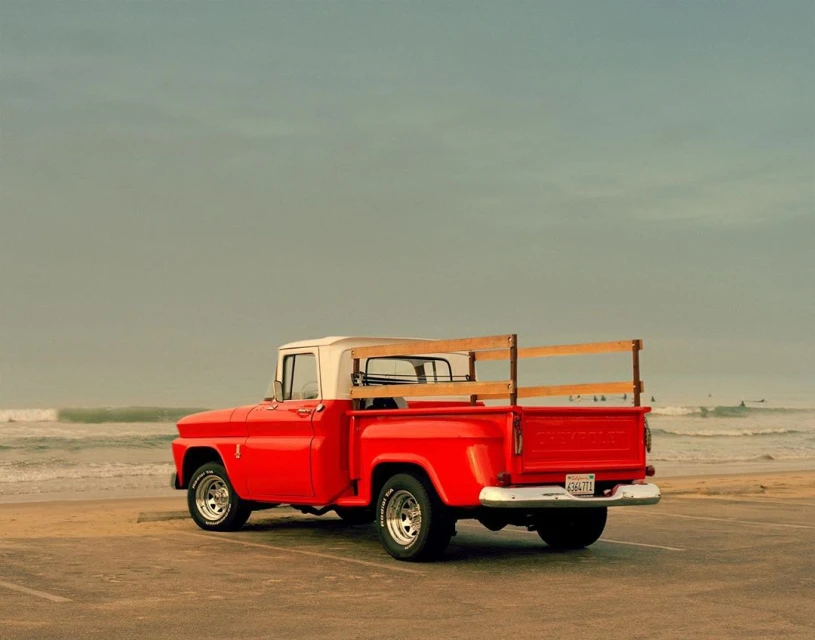 A classic red and white Chevrolet pickup truck parked on a beach, waves and surfers visible in the background, under a cloudy sky.
