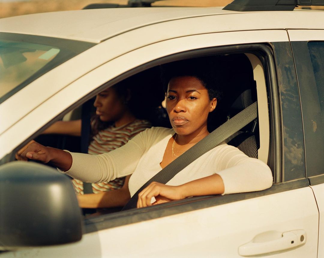 Two African-American women are inside of a white SUV in an outdoor desert environment during the daytime, captured in a medium shot.