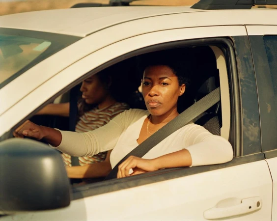 Two African-American women are inside of a white SUV in an outdoor desert environment during the daytime, captured in a medium shot.