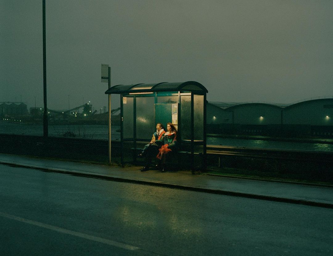 Two people sit at a bus stop on an overcast day with industrial buildings visible across the waterway in the background.