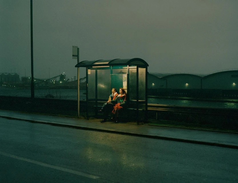 Two people sit at a bus stop on an overcast day with industrial buildings visible across the waterway in the background.