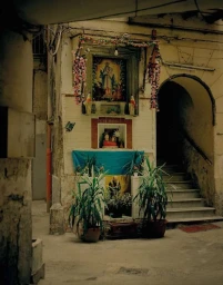 A roadside shrine with framed religious imagery, floral garlands, and potted plants set against a weathered stone wall