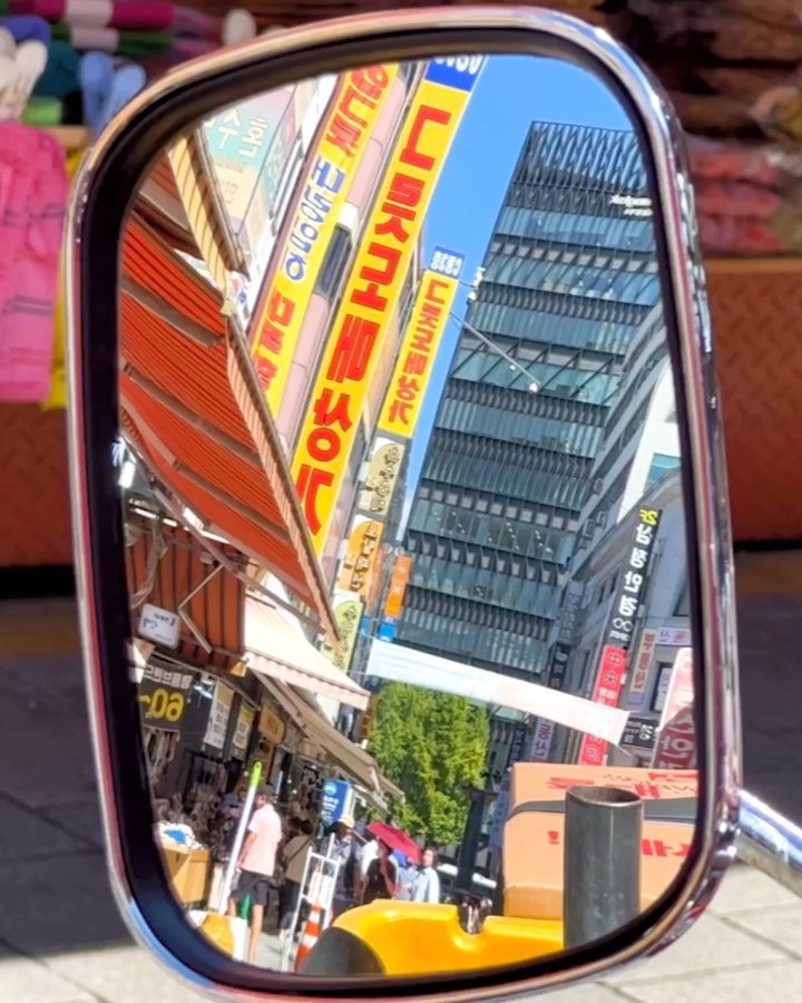 Reflection in side mirror shows a bustling city street with pedestrians and tall buildings under a clear blue sky, on a sunny day.
