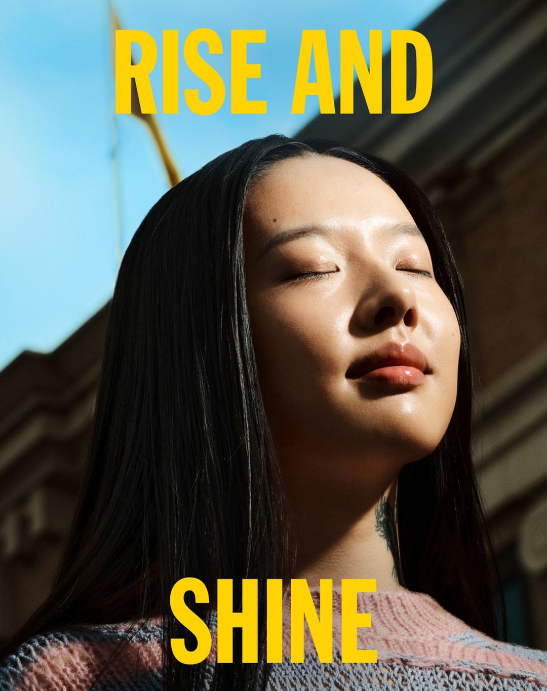 Close-up of a serene woman with closed eyes outdoors with a building behind her. Yellow text above her reads 'Rise and Shine'.