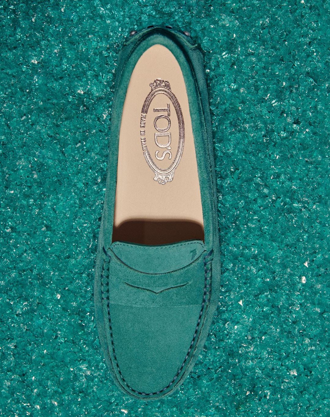 Overhead studio shot of a green Tod's loafer shoe displayed on a decorative green surface with crystal-like pieces.