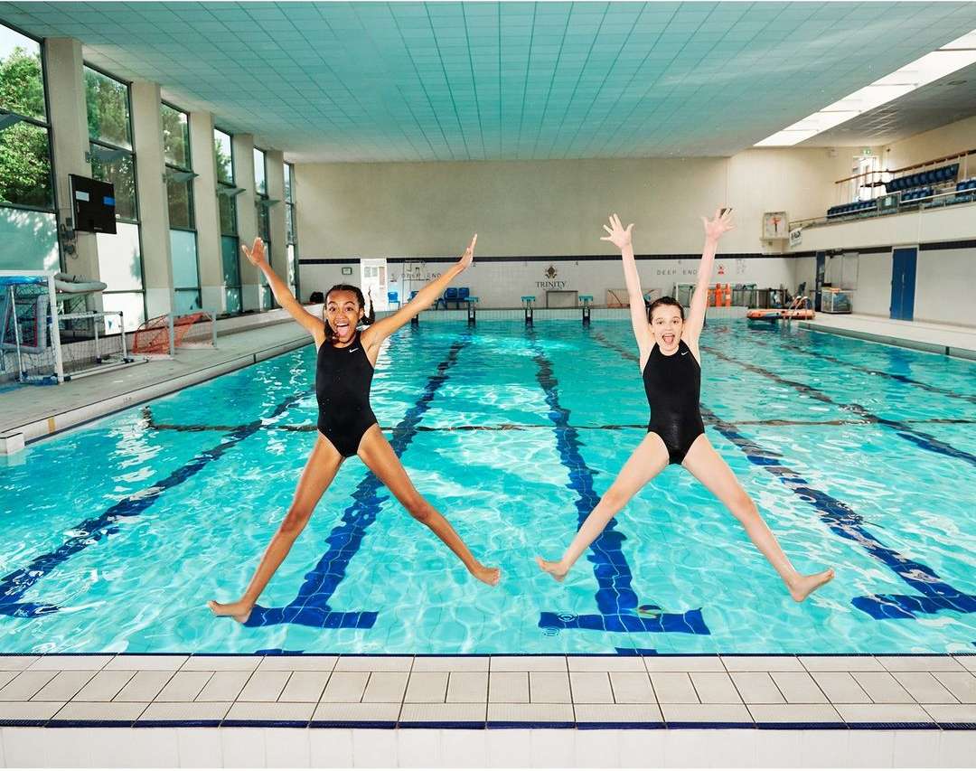 Two young girls in black swimsuits jumping mid-air with arms raised over a bright blue indoor swimming pool, tile floors, and natural light.