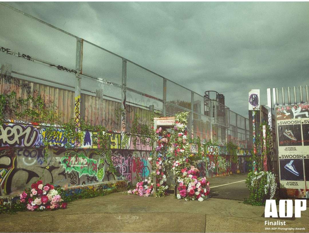Urban landscape featuring graffiti-covered walls, chain-link fence, and floral arrangements, photographed for the AOP Photography Awards.