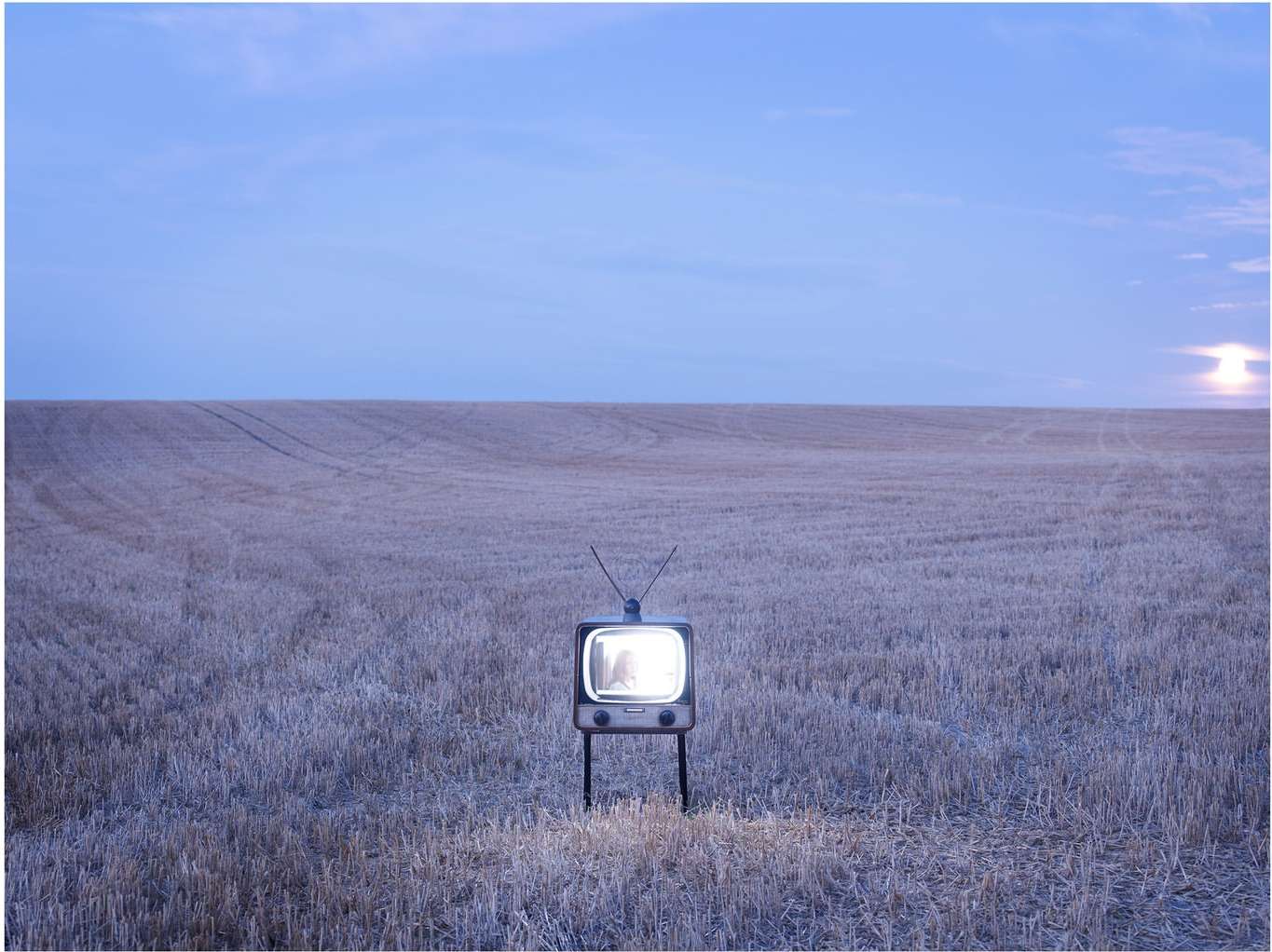 Vintage TV set glowing in a rural field against a light blue sky at dusk, creating a surreal, illuminated contrast.