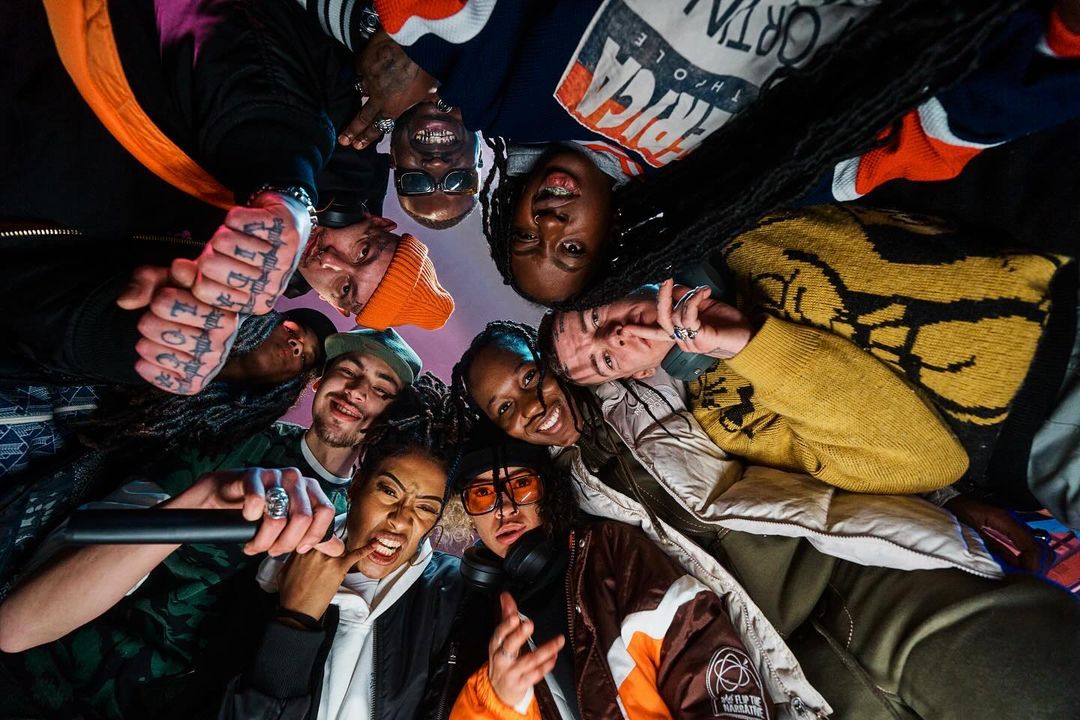 Group of diverse young adults huddle together in an eye-level low angle shot, wearing trendy streetwear in an indoor setting.