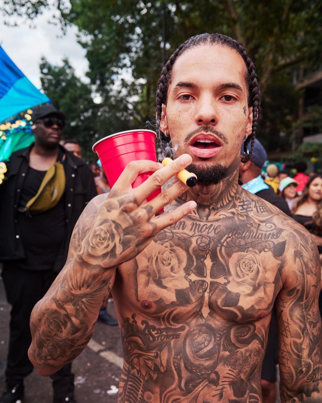 Tattooed man poses holding a drink and smoking, with crowd and trees blurred in background, captured in a medium shot.