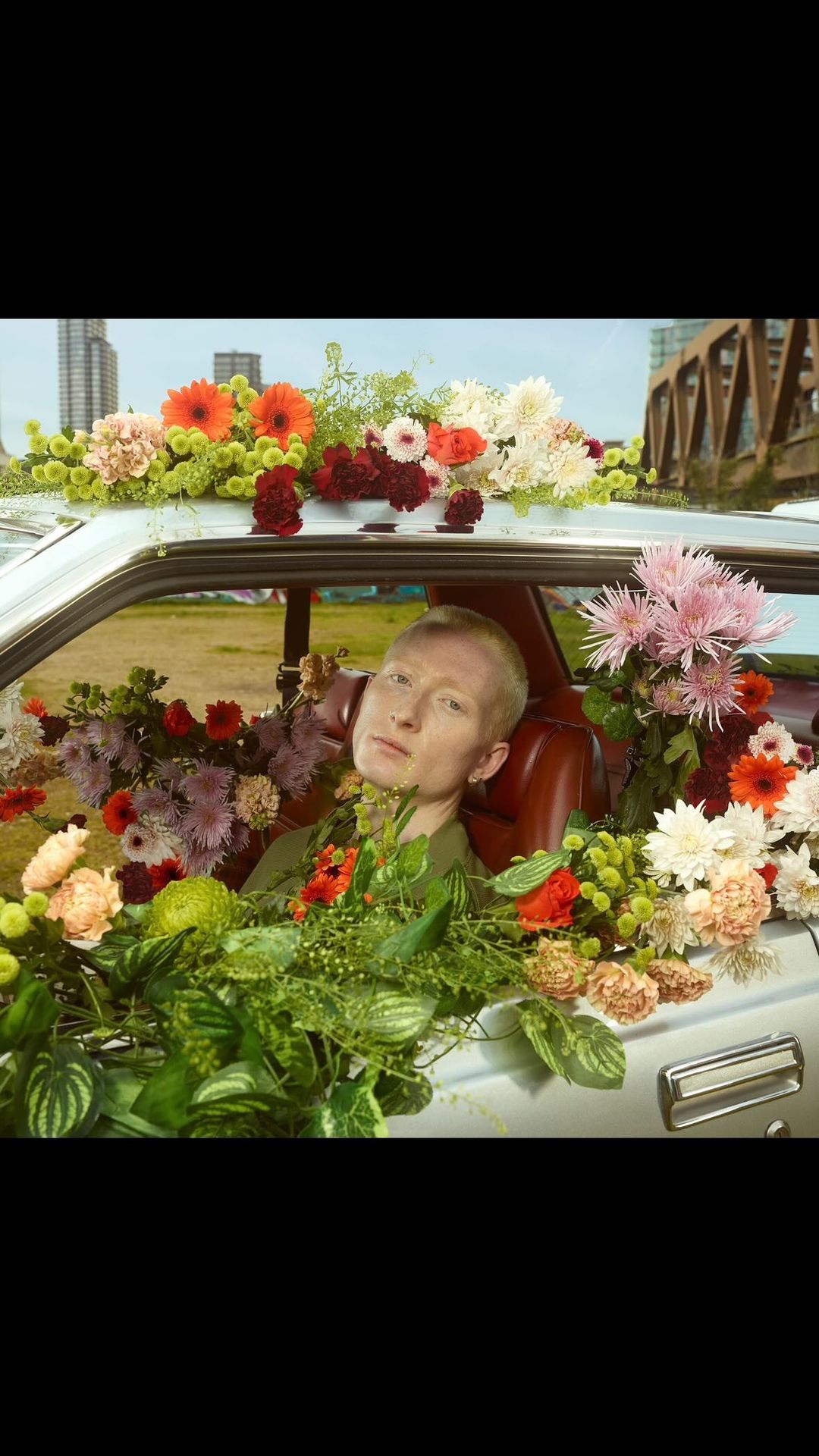Person sits in a vintage car filled with various flowers; city buildings and industrial structures appear in the background.