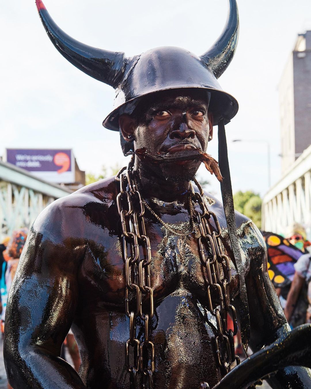Carnival performer covered in black paint, wearing horned helmet and large chain necklace, poses outdoors with a serious expression.