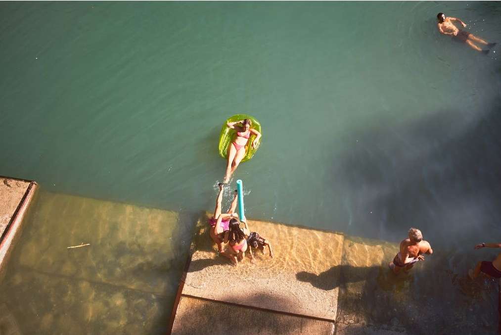 Aerial view of two women in bikinis near the water's edge, one on a raft, with other swimmers in the turquoise water.