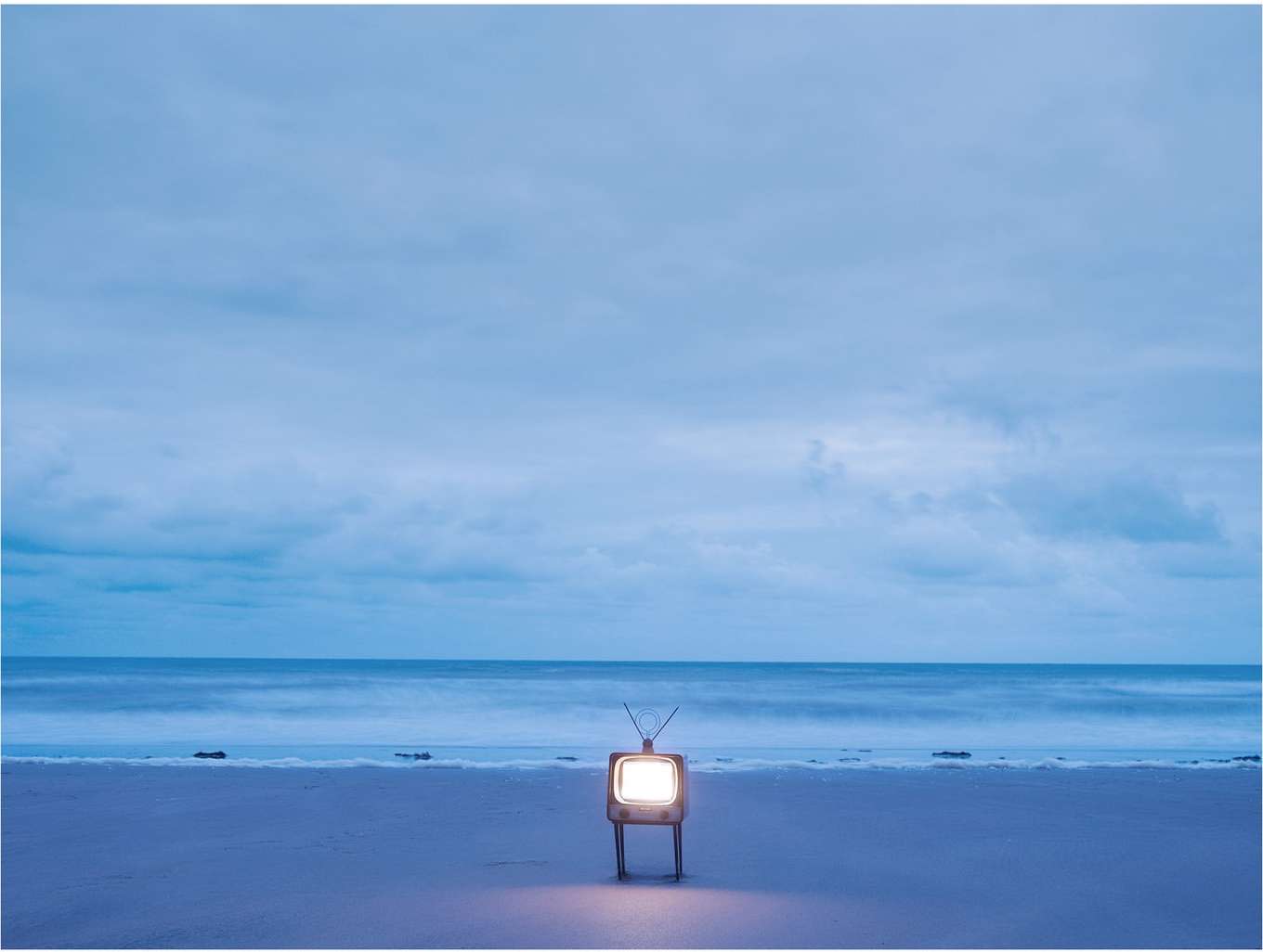 Vintage television set with illuminated screen on a sandy beach against a backdrop of soft blue ocean and cloudy sky.