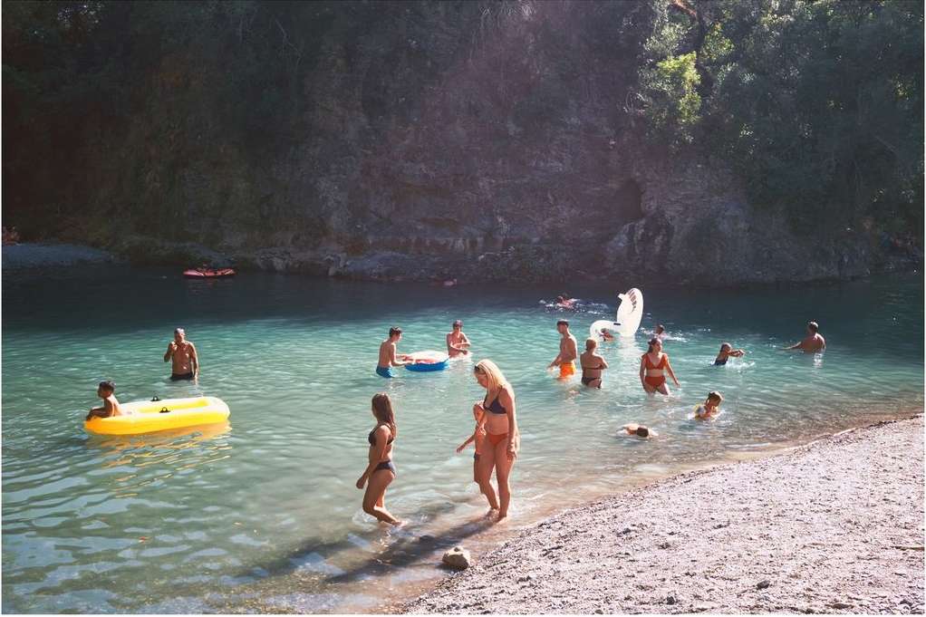 Group of people swimming in a turquoise river surrounded by a rocky hillside and lush trees on a sunny day in a natural setting.