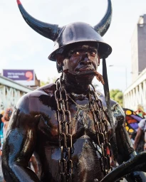 Black man wearing a horned helmet and heavy metal chains with a dark glossy body paint coating outdoors in London.