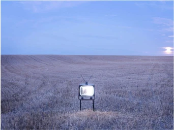A vintage television set stands upright in the middle of a vast harvested field under an evening sky.