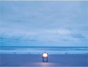 A vintage television set stands upright on a deserted sandy beach with its screen glowing brightly against a pale blue overcast sky and ocean horizon.