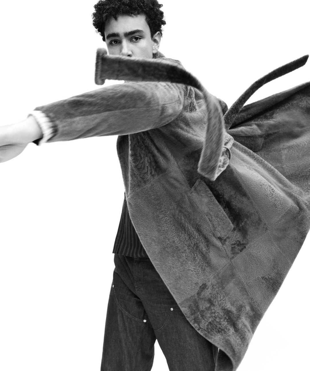 Monochrome fashion shot of a young adult male with curly hair wearing an oversized wool coat looking at the camera against a white backdrop.