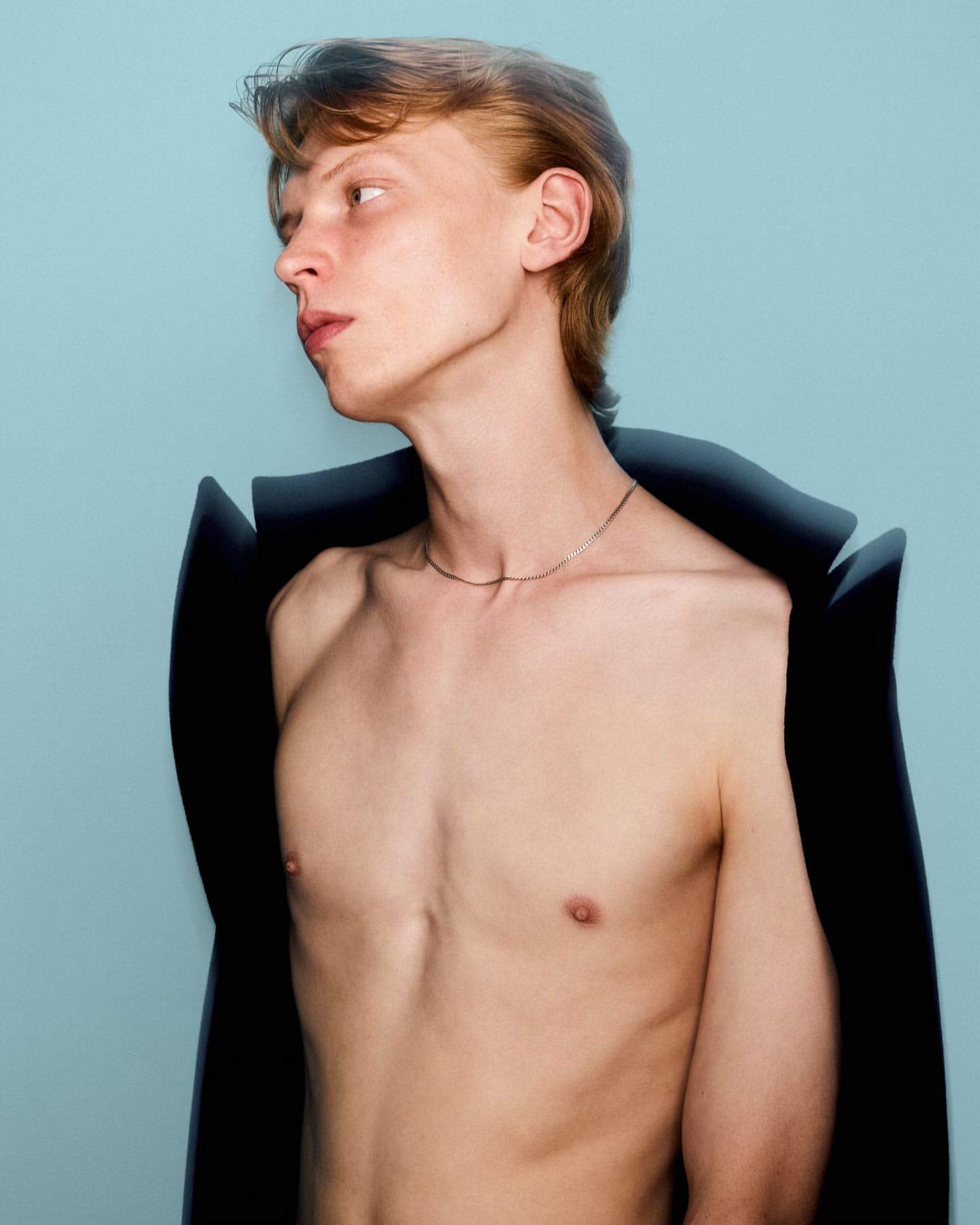 Portrait of shirtless fair-skinned adult male with reddish hair and silver necklace on a light blue background, wearing black material.