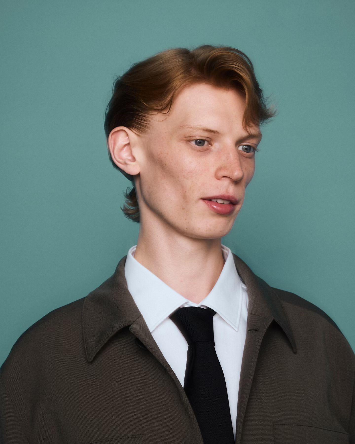 Portrait of a redhead adult male in a white collared shirt and black tie, wearing a brown jacket against a turquoise background.