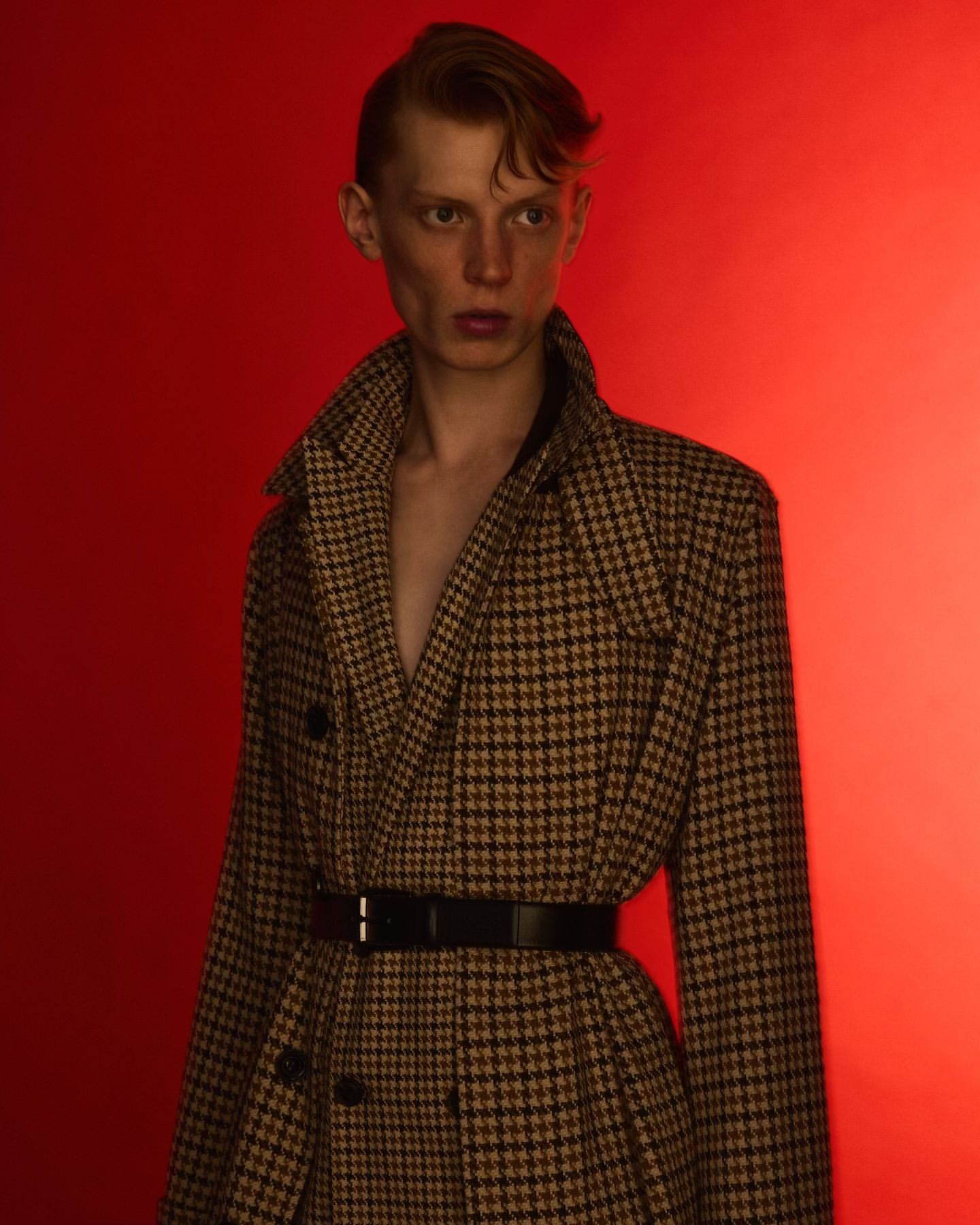 Adult male model in a houndstooth pattern coat with a black belt posing against a red background in a fashion photoshoot.