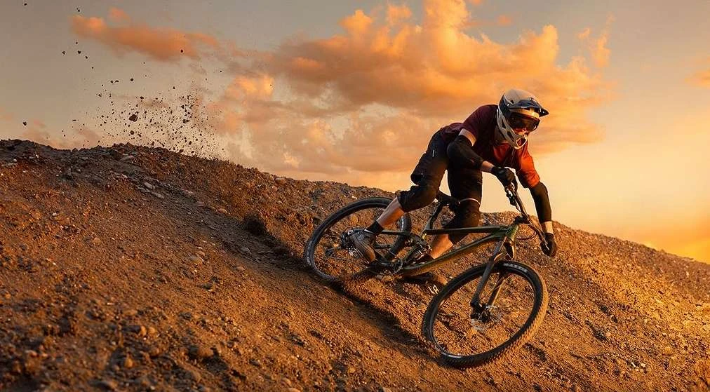 A mountain biker rides downhill on a dirt track with a golden sunset sky. The rider is wearing a helmet and protective gear.